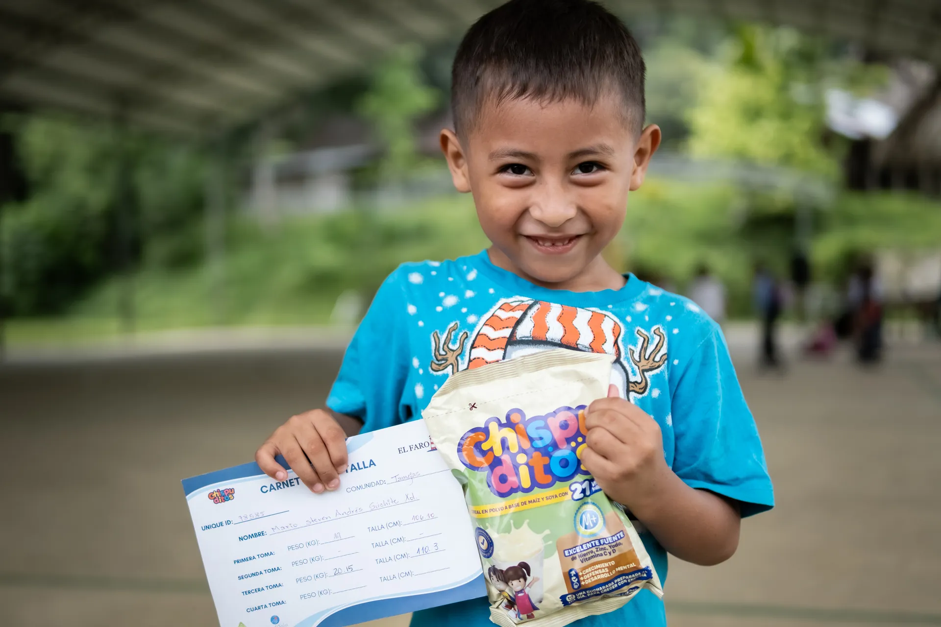 A child smiles while holding materials from an El Faro nutrition program.
